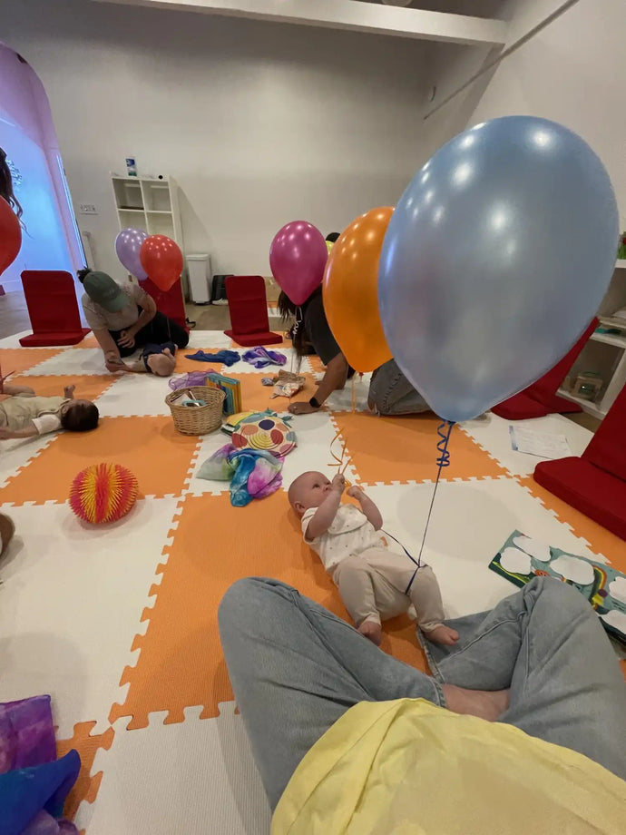 Baby lying on colorful foam floor mats surrounded by balloons and toys.